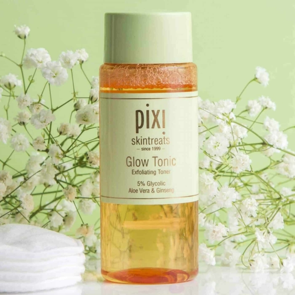 Sur un fond vert clair orné de délicates fleurs blanches, un flacon de PIXI Glow Tonic Lotion Tonique Exfoliante 100 ml occupe le devant de la scène. L'étiquette accentue la présence d'acide glycolique à 5 %, d'aloe vera et de ginseng. Des disques de coton sont élégamment empilés à proximité. Dakar