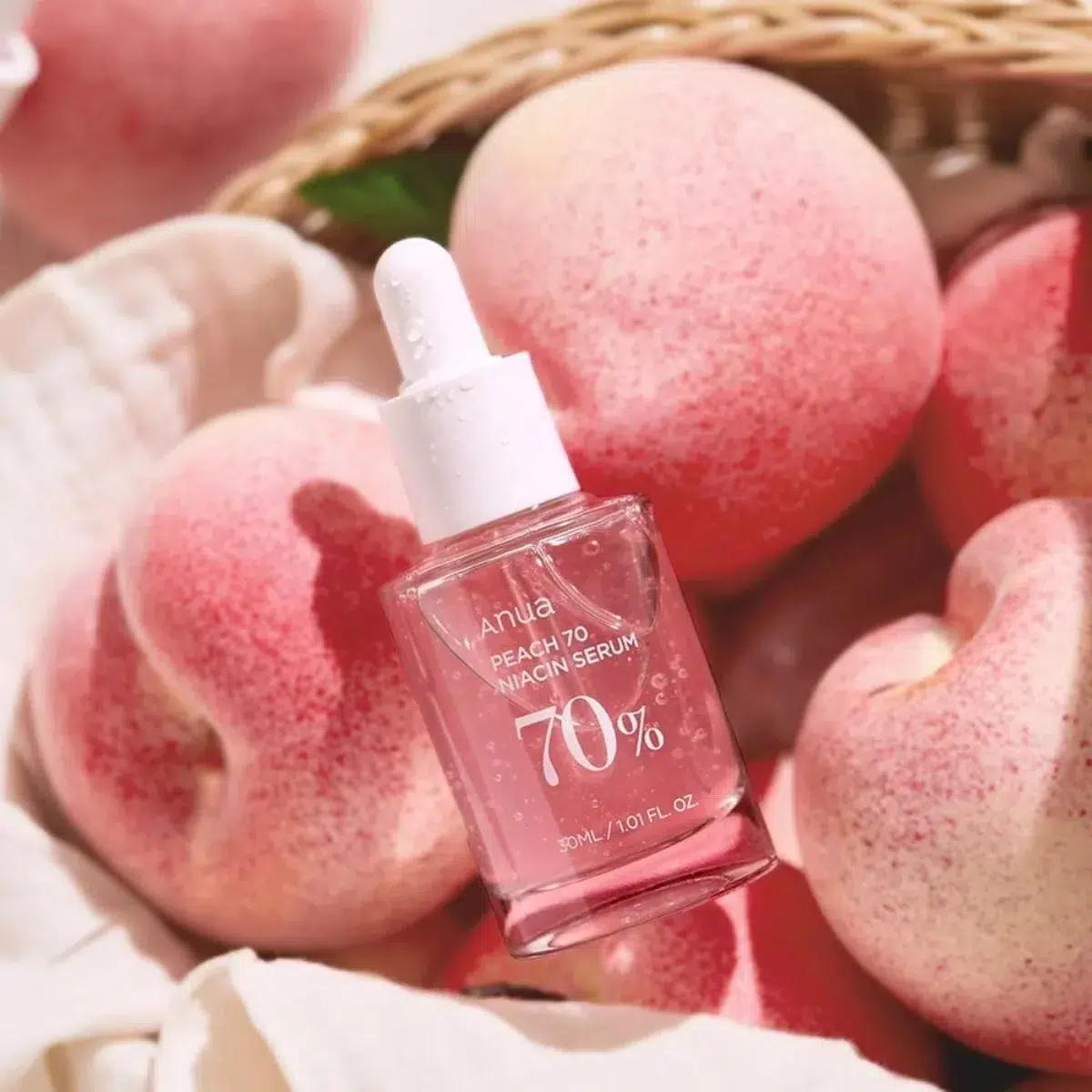 Flacon compte-gouttes de ANUA Peach 70% Niacinamide Sérum Éclat Peau de Pêche 30 ml sur un panier de pêches fraîches avec un chiffon doux à côté.