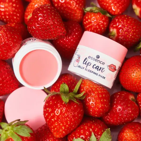 Un pot ouvert de Gelée de Masque à Lèvres 02 Milkshake à la Fraise Essence Cosmetics 8 g repose à Dakar, au Sénégal, au milieu de fraises fraîches. Le couvercle rose du masque pour les lèvres et son étiquette ornée d'illustrations de fraises mettent magnifiquement en valeur les fraises rouges vives éparpillées tout autour. Dakar