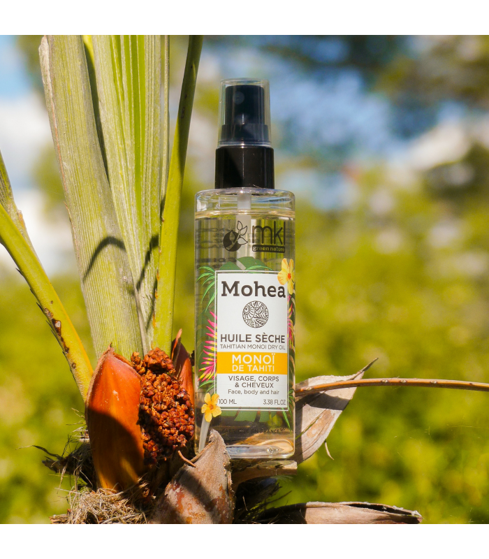 Un flacon de MKL Huile sèche - Monoï - 100 ml est niché au milieu d'un feuillage tropical avec un fond naturel flou