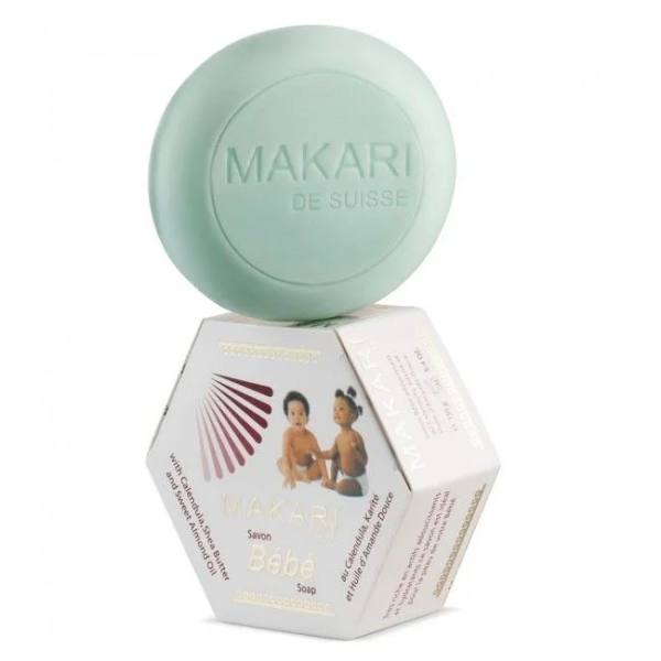 Un savon rond vert étiqueté « Makari de Suisse » repose sur une boîte hexagonale indiquant « Makari Bébé Savon au Calendula et Beurre de Karité 155 g »