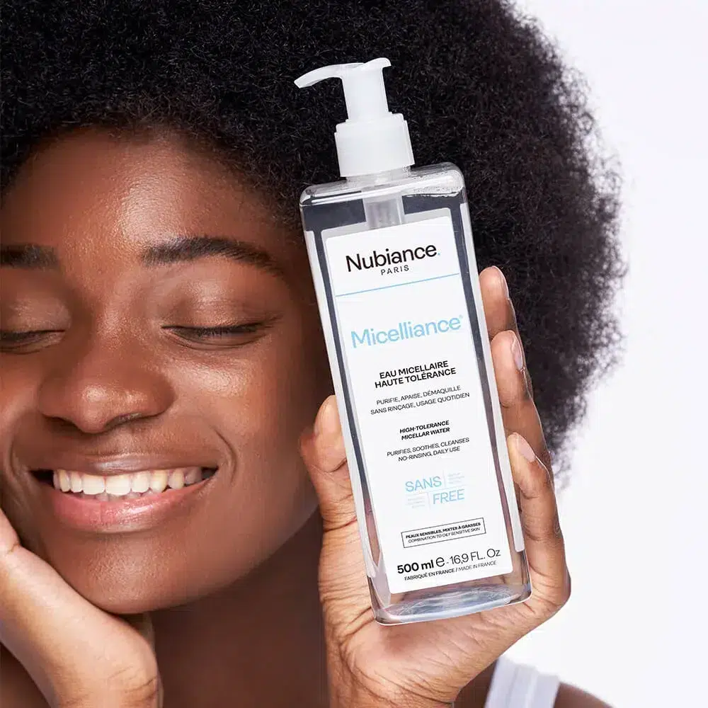 Une personne souriante aux cheveux bouclés tient un flacon d'eau nettoyante Nubiance Micelliance Eau Micellaire Haute Tolérance Peaux Sensibles à Grasses 500 ml contre sa joue. dakar