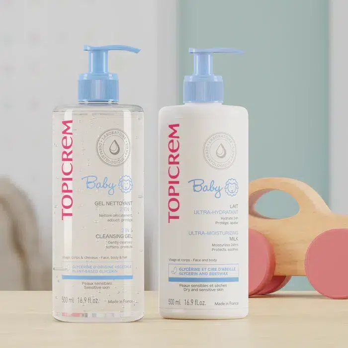 Deux flacons de Topicrem Kit Bébé Nourrissant Peaux Sensibles Lait + Gel Nettoyant 2en1 sont élégamment présentés sur une surface en bois