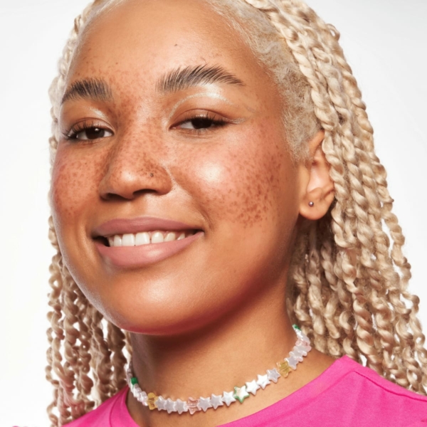 Une personne aux cheveux blonds tressés et aux taches de rousseur sourit à la caméra. Elle porte un haut rose, probablement rehaussé par le Blush Liquide Baby Got Blush d'Essence Cosmetics, et un collier avec des perles en forme d'étoile sur un fond blanc uni. Dakar