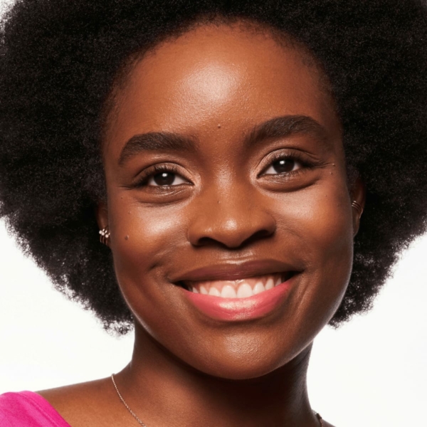 Une personne avec une coupe de cheveux afro, portant un haut rose et de petites boucles d'oreilles, sourit directement à la caméra. Elle présente le Blush Liquide Baby Got Blush d'Essence Cosmetics sur un fond blanc uni. Dakar