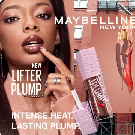 Une femme aux lèvres brillantes touche son menton, mettant en valeur le nouveau gloss Lifter Plump de Maybelline. La publicité présente une image du gloss et un piment rouge ardent, avec un texte mettant l'accent sur « chaleur intense » et « volume durable ». Dakar